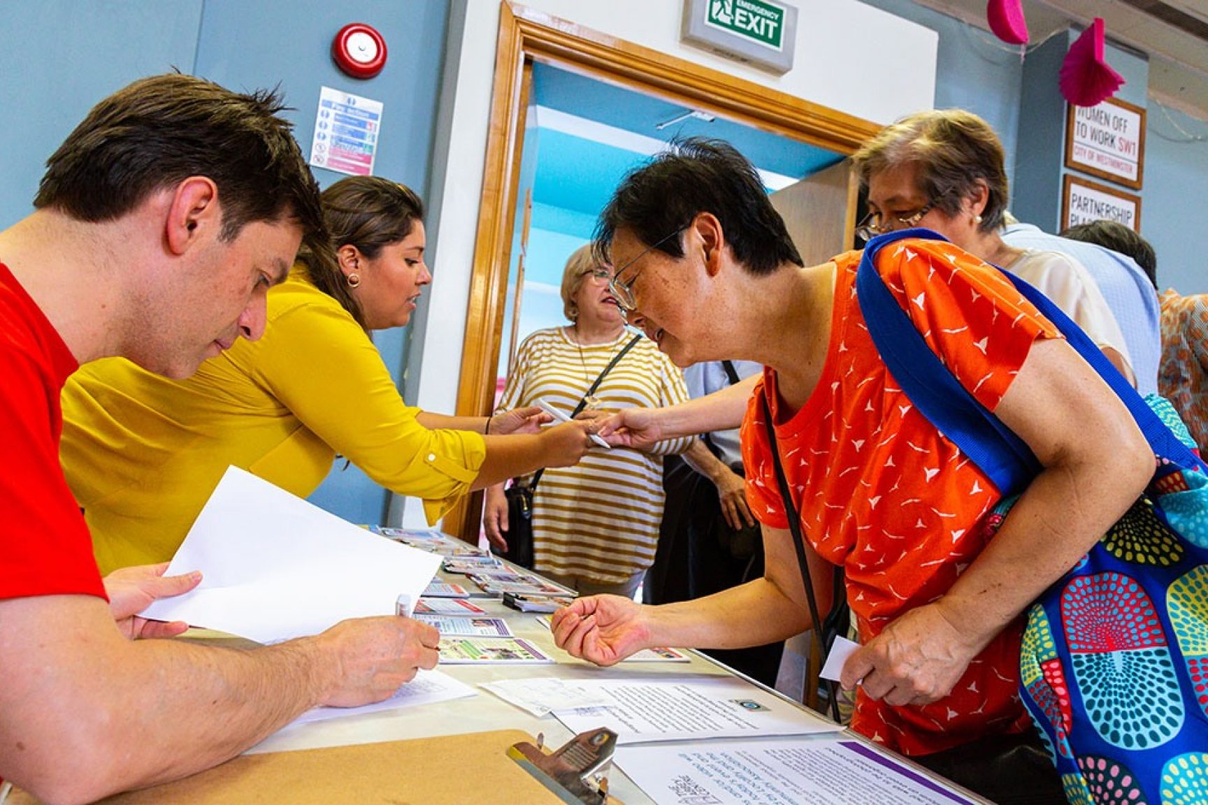 Registration at an event. Two people are signing in attendees, everyone is wearing brightly coloured clothing.