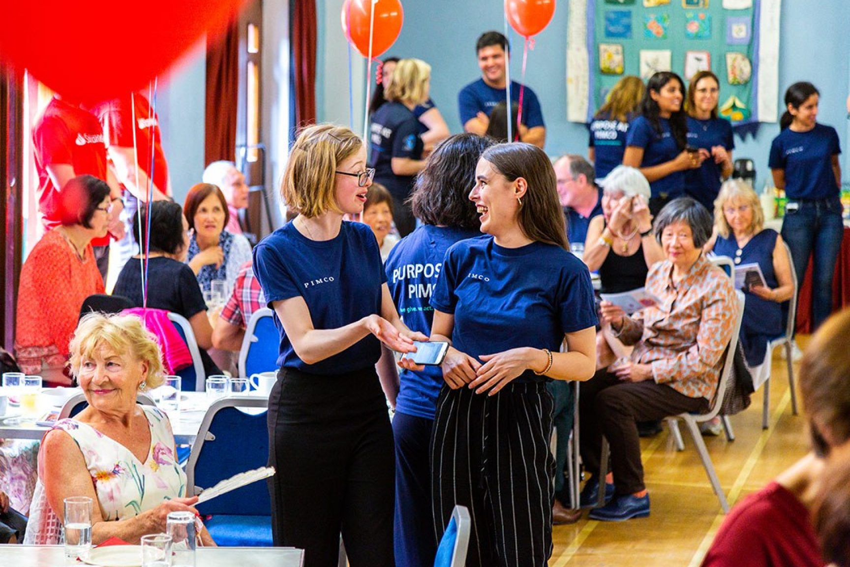 A community event, there are balloons, people seated at tables having a nice time. There a two volunteers chatting and smiling at each other in the middle of the photo.