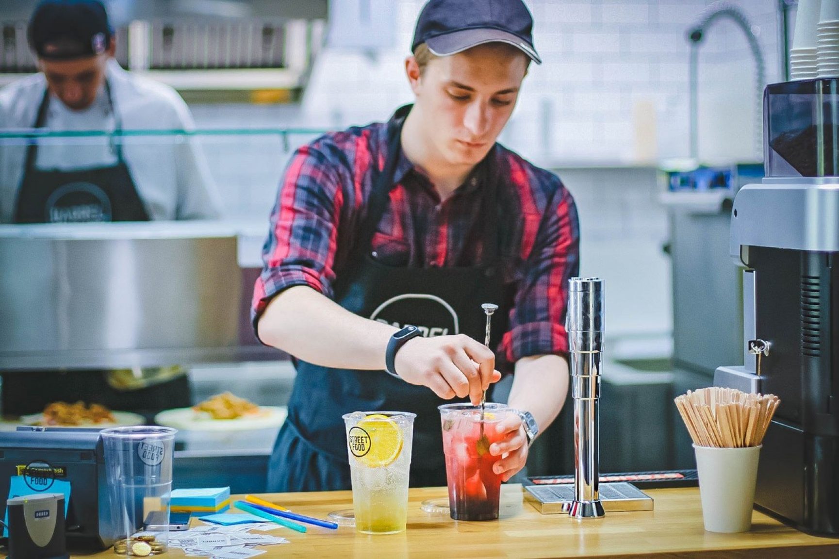 A person mixing drinks behind a counter. He is wearing a cap and an apron.