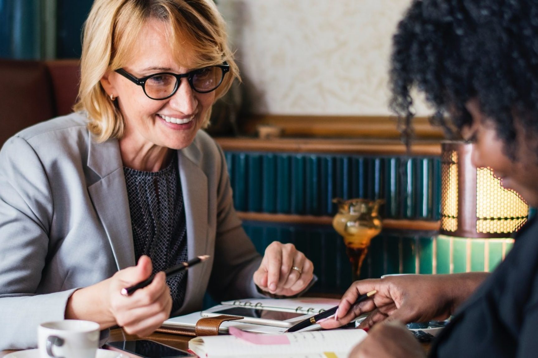 Two women in suits, they are smiling and discussing something. They each have books and diaries in front of them at a table.