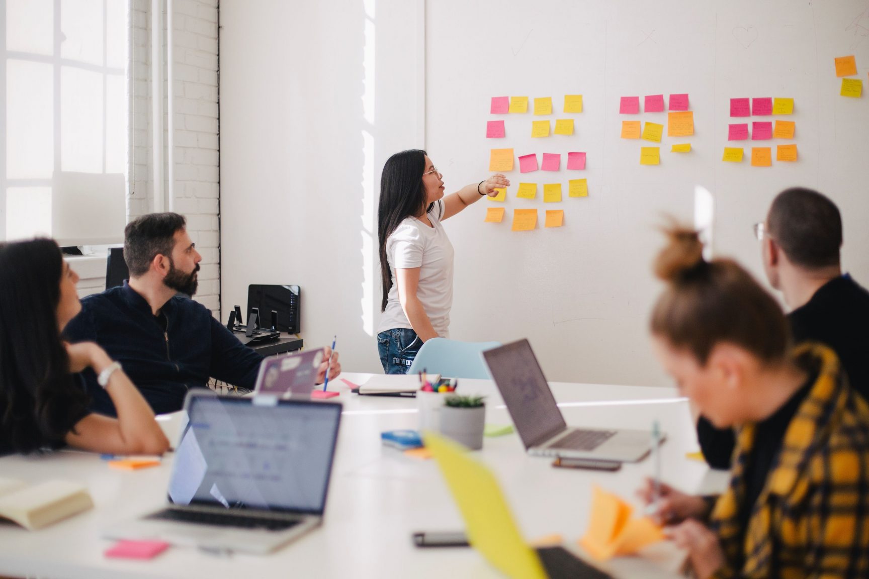 A group of people at work. They are in a meeting room, and have lots of yellow and pink post-its on the far wall.